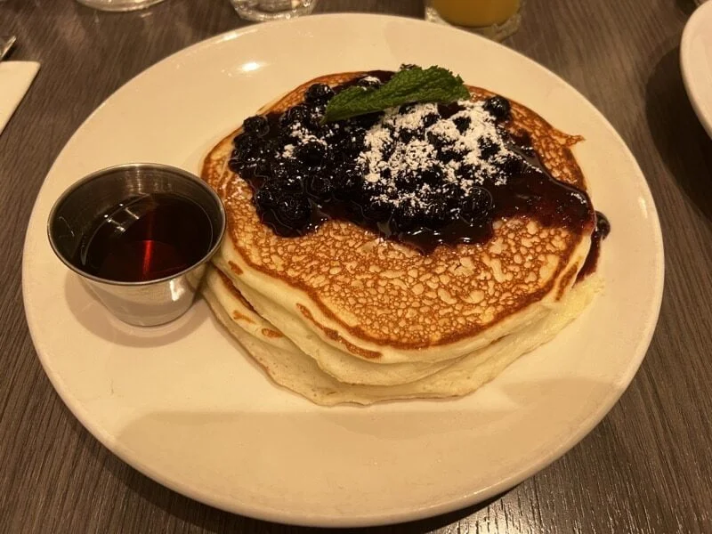 stack of glutenfree pancakes with blueberries on top, and silver pot of syrup ontop of white plate